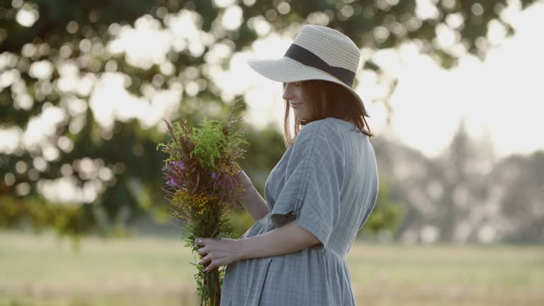 Pregnant woman in linen dress and hat walks with wild flowers bouquet outdoors by oak tree in meadow on sunset