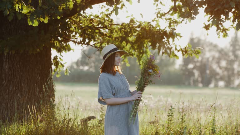 Pregnant woman in linen dress standing with flowers bouquet outdoors by oak tree in the meadow during sunset