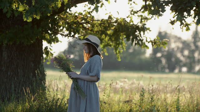Pregnant woman in linen dress and hat walks with wild flowers bouquet outdoors by oak tree in meadow on sunset
