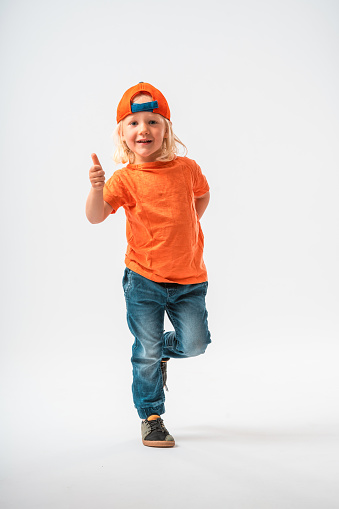 Caucasian boy of kindergarten age wearing a  shirt and a baseball cap back to front. Standing on one foot, other one bent behind the knee, showing thumbs up. Looking at the camera and smiling, full length image.