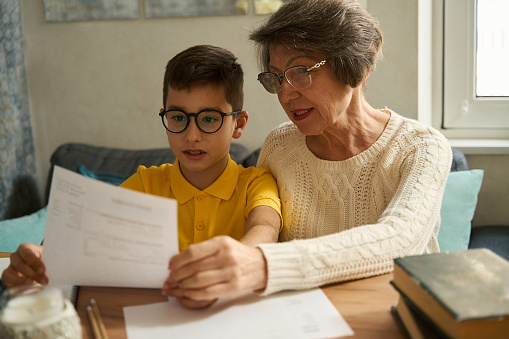 Kid sitting at the table with granny, holding paper, reading, doing school homework at home