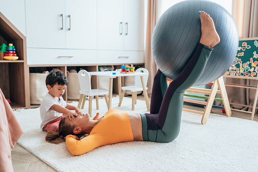 Woman working out with her toddler son around. Doing exercises with fitness ball at home. Young woman exercising in child's room. Fit mom doing exercises