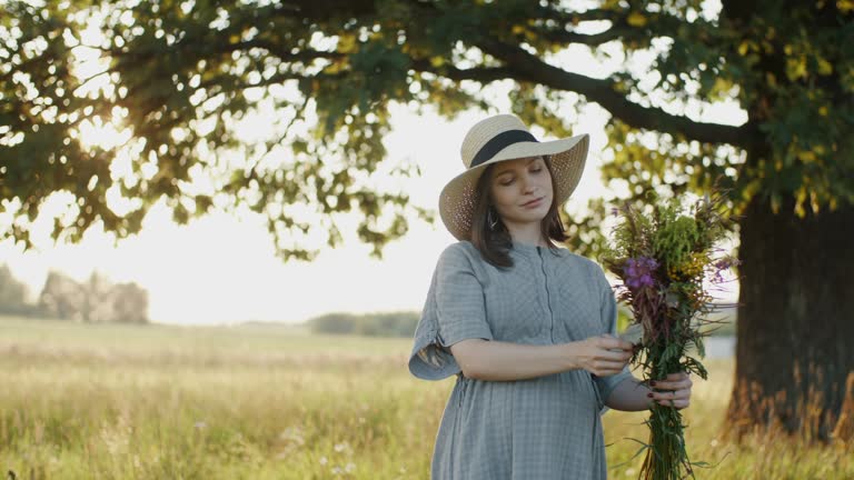 Pregnant woman standing outdoors with flower bouquet by oak tree in meadow during sunset