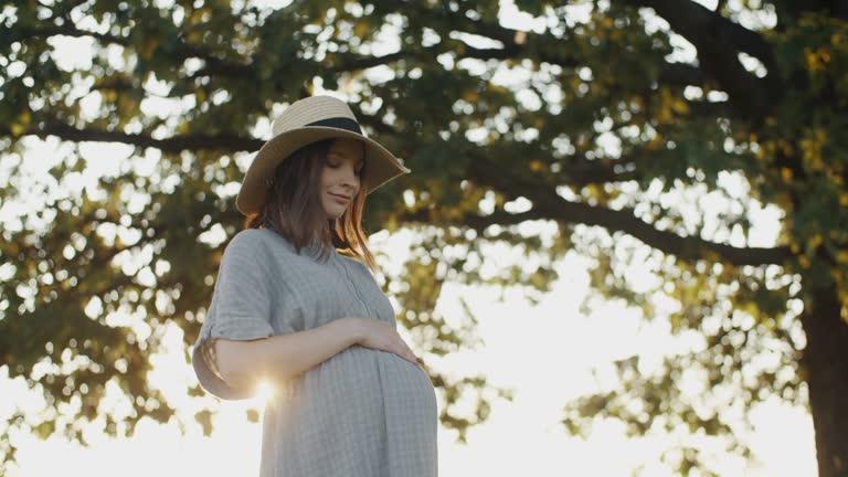 Cute pregnant woman wearing straw hat stroking belly standing by oak tree in meadow during sunset