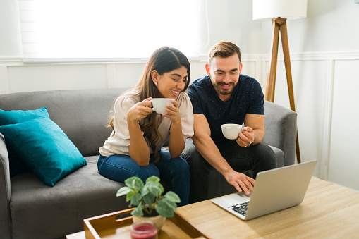 Cheerful young woman and man drinking coffee while using the laptop for online shopping
