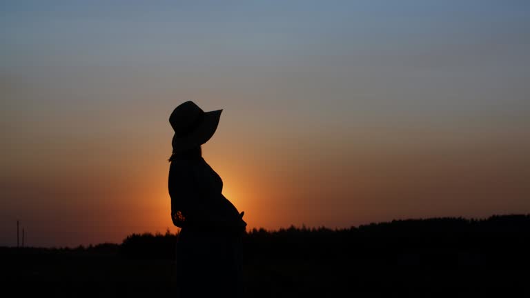 Silhouette pregnant woman in dress and hat touching belly standing in meadow outdoors during sunset