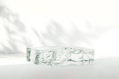 Natural ice form transparent acrylic glass podium on white table counter in dappled sunlight, leaf shadow on wall background for luxury beauty, cosmetic, organic, health, food and drink product display backdrop