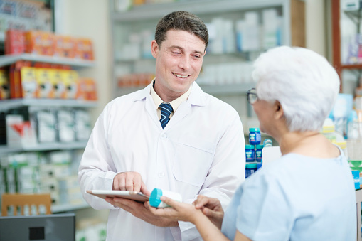 Smart male pharmacist at the drugstore wearing white gown talking, giving advice, explaining, suggesting, and recommending to client or patient about the prescription and medications. Medicine and healthcare concept.