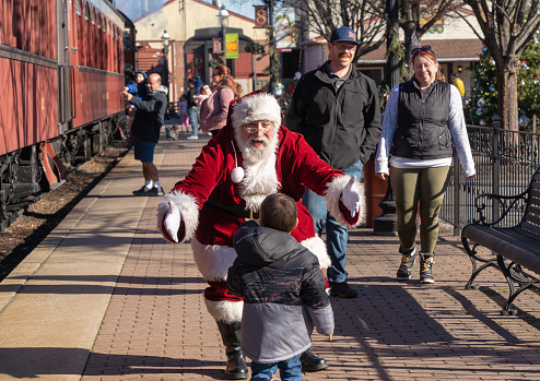 December 11, 2022: Strasburg, Pennsylvania: Parents watch as Senior Gentlemen dressed as Santa Claus greets small child at Strasburg Railroad Christmas train ride experience.