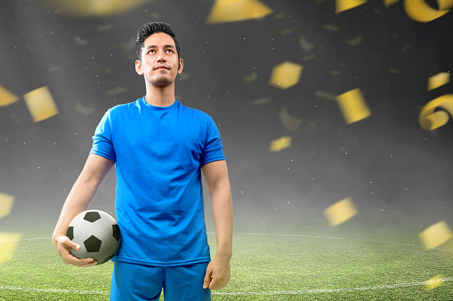 An Asian football player man in a blue jersey is standing and holding the ball on the football field