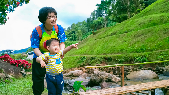Happy Asian aunt with her little grandson enjoying nature beside a small stream with bamboo bridge on green hill.