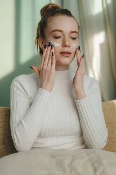 Woman with closed-eyes doing routine skin care at home applying cream on her face while sitting on sofa at home. Caucasian girl taking care of herself