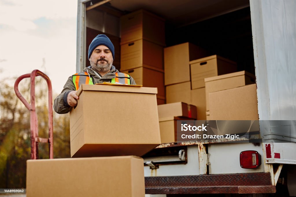 Male worker unloading cardboard boxes from delivery van. Mid adult courier loading delivery boxes for shipment into a truck. Moving Van Stock Photo