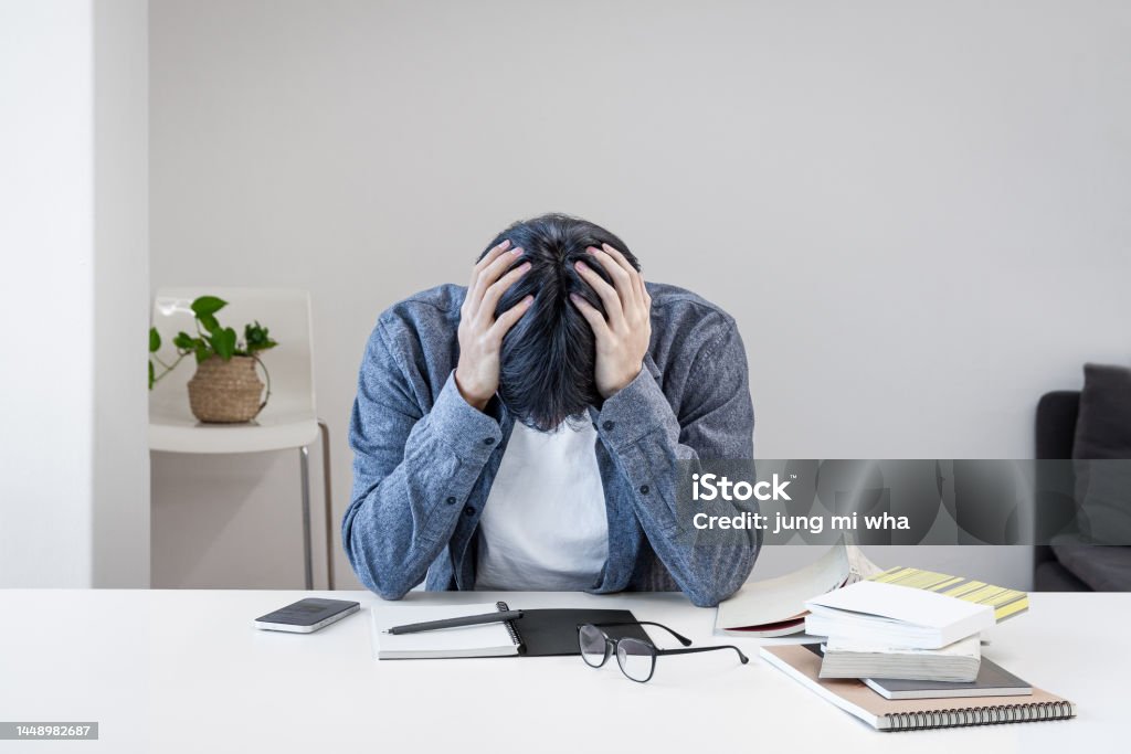 A troubled Asian Korean college student is holding his head in his hand because he is stressed out in the library. There are books, notebooks, pens, glasses, cell phones, etc. on the desk. Student Stock Photo