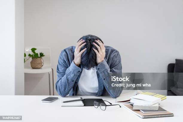 A Troubled Asian Korean College Student Is Holding His Head In His Hand Because He Is Stressed Out In The Library There Are Books Notebooks Pens Glasses Cell Phones Etc On The Desk Stock Photo - Download Image Now