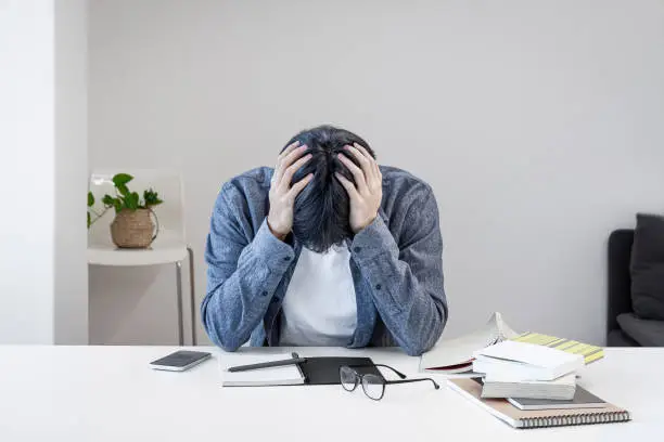 Photo of A troubled Asian Korean college student is holding his head in his hand because he is stressed out in the library. There are books, notebooks, pens, glasses, cell phones, etc. on the desk.