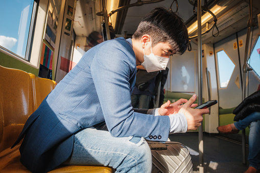Asian businessman passenger in suit wearing protective face mask using mobile phone and sitting inside BTS sky train or MRT underground train at railway platform while traveling to work, Bangkok, Thailand, Transportation and lifestyle concept