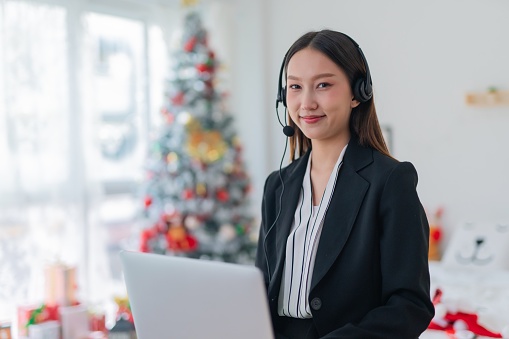 Portrait photo of a young beautiful smart asian lady businesswoman working on laptop and providing customer support assistance over her wired microphone headset during Christmas holidays. Background is a nicely decorated Christmas tree.