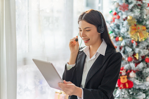 Portrait photo of a young beautiful smart asian lady businesswoman working on digital tabletand providing customer support assistance over her wired microphone headset during Christmas holidays. Background is a nicely decorated Christmas tree.