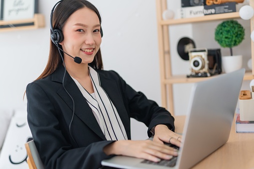 Portrait photo of a young beautiful smart asian lady customer support agent representative working on laptop and providing customer support assistance over her wired microphone headset in a random cafe corner