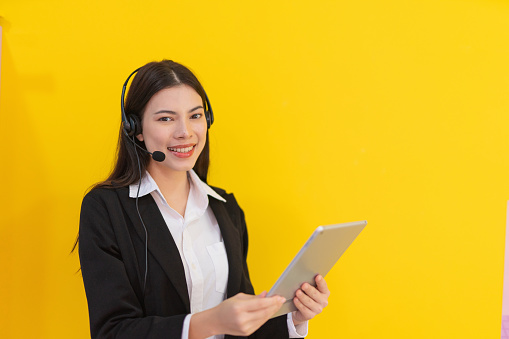 Portrait photo of a young beautiful smart asian lady customer support agent representative with a digital tablet and her wired microphone headset on a yellow color background