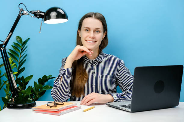 青の背景に、オフィスの机に座って仕事をし、ノートパソコンの小道具をあごで、カメラを見て幸せに見える、成功した笑顔の従業員のビジネスマンのポートレート。達成キャリアコンセプ� - looking at camera smiling desk isolated ストックフォトと画像