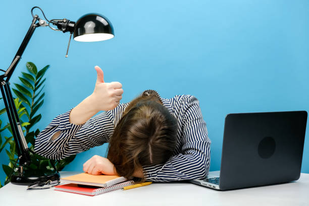 stanca segretaria esausta dipendente donna d'affari in camicia seduta lavoro sonno posato la testa giù sulla scrivania dell'ufficio con pc portatile mostra il gesto del pollice in su, isolato sulla parete di sfondo di colore blu in studio - exhaustion foto e immagini stock