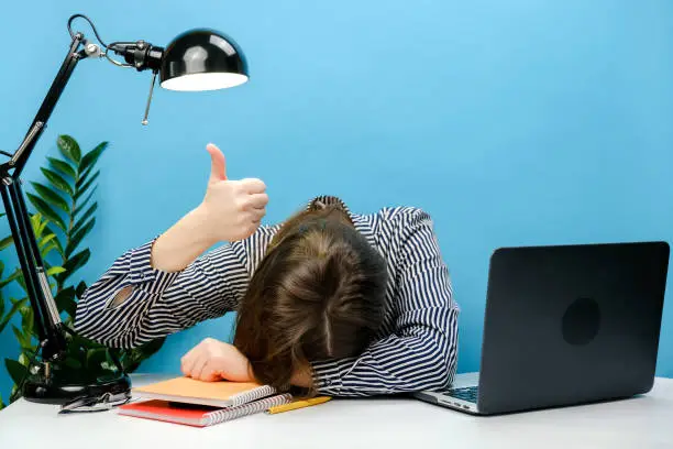 Photo of Tired exhausted secretary employee business woman in shirt sitting work sleep laid her head down on office desk with pc laptop show thumb up gesture, isolated on blue color background wall in studio