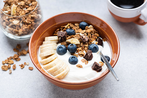 Yogurt with granola, banana, blueberries and dark chocolate in bowl, grey cocnrete table background, closeup view. Healthy food