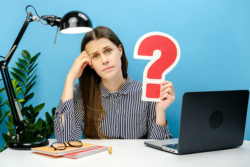 Pensive young employee female 20s in shirt sitting work at white office desk pc laptop holding red question mark sign, posing isolated over blue color background wall in studio. Business concept