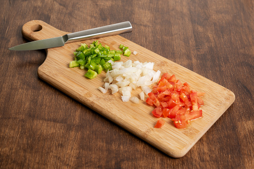 Tomato, pepper and onion cut into small pieces on a cutting board with a knife.