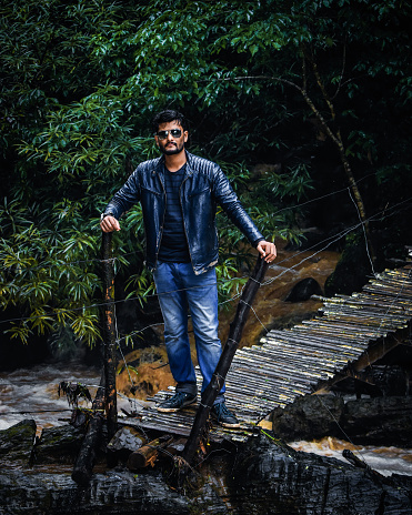 Handsome guy wearing jacket and sunglasses standing alone at forest over a wet wooden bridge.(solo traveler concept)