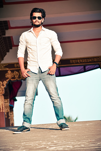 Handsome fit model with beard and sunglasses, looking at camera, with hands in pocket.