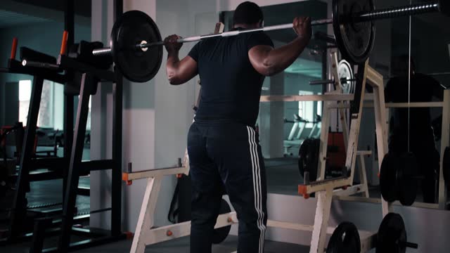 Muscular man training with barbell, doing back squats in gym, back view. Black Bodybuilder training in gym with barbell.Athlete workout doing exercise for legs indoors.