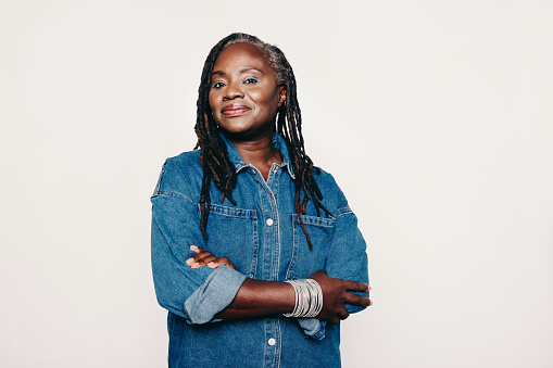 Stylish mature woman looking at the camera while standing against a grey background with her arms crossed. Fashionable woman with dreadlocks wearing a denim jacket and make-up.