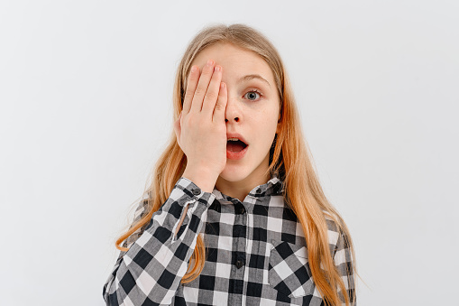 Surprised teen girl making facepalm, looks amazed open mouth and covers half of face with palm, looks with one eye at camera, stands over white studio background