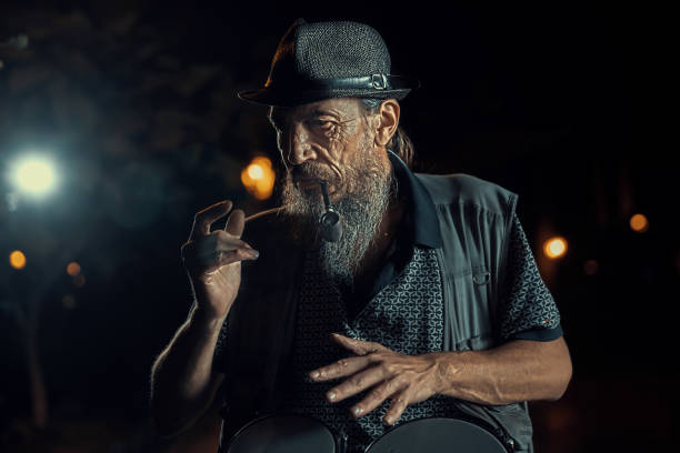 istock Old angry bearded drummer in a hat smokes a pipe at night street 1449029248