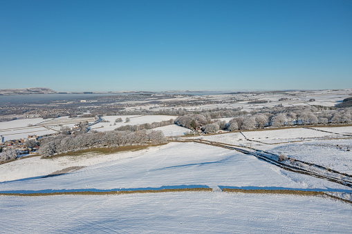 Aerial drone photo of the town of Mereclough in the town of Burnley in Lancashire, England showing the farmers fields on a snowy winters day in the UK with snow covering the fields and a blue sky.