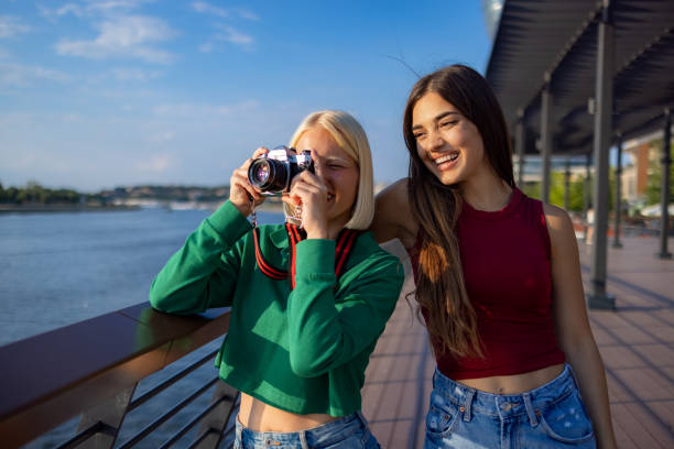 Happy female friends enjoying in spring day on a pier by the river while one of them is photographing with digital camera.