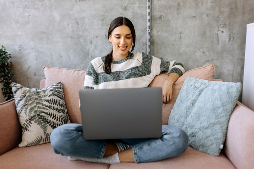 Smiling young woman using laptop, sitting on couch at home
