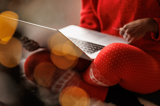 Selective focus shot of unrecognizable young Black woman sitting on the window sill, holding her credit card, doing online Christmas shopping via laptop. Christmas tree glistening in the blurred foreground.