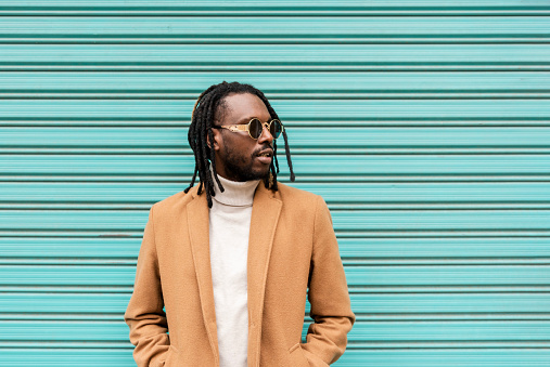 handsome african american man with braids wearing stylish sunglasses looking away while standing against blue wall