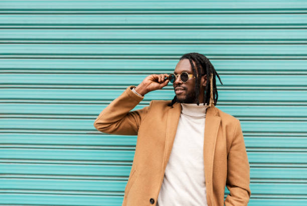 istock smiling african american man with braids adjusting stylish sunglasses looking away while standing against blue wall 1449034799