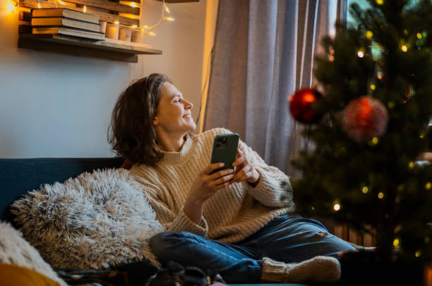 istock Young cheerful happy woman using smartphone while sitting at home on sofa next to christmas tree, Christmas and New Year holidays concept 1449034840