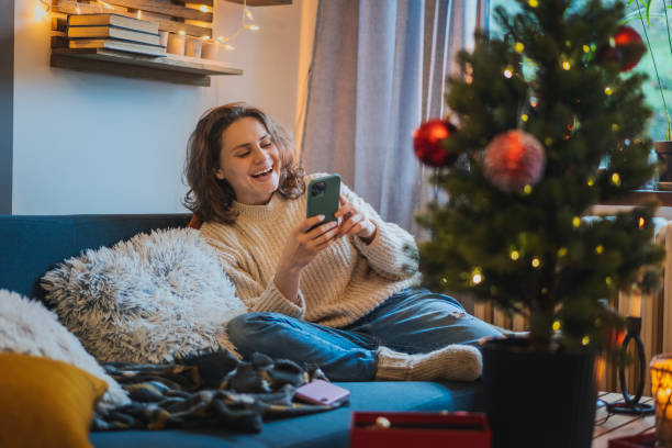 istock Young cheerful happy woman using smartphone while sitting at home on sofa next to christmas tree, Christmas and New Year holidays concept 1449034877