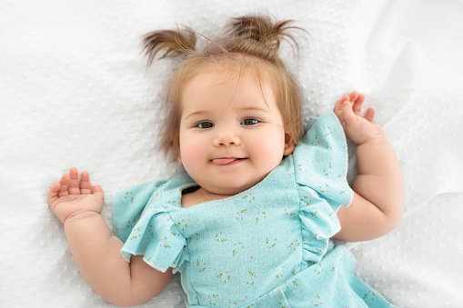 Close up happy baby girl showing tongue and looking at camera. Cute child with pigtails in white bedroom lying on bed. Childhood, babyhood, people concept.