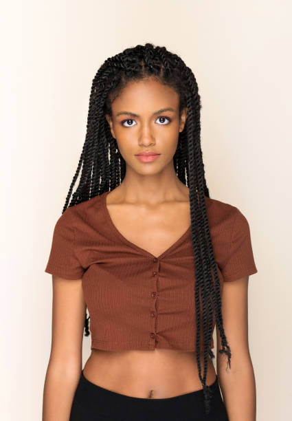 Vertical portrait of young black woman with Afro braids in brown top standing on beige background looking at camera