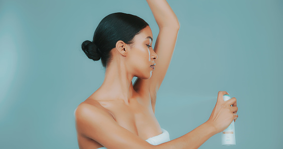 Studio shot of beautiful conscious young woman wearing a towel spraying deodorant on her underarms against blue background