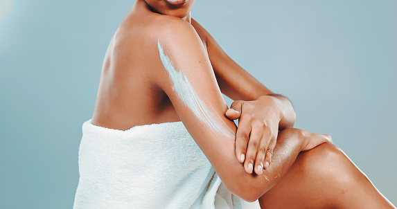 Midsection of African American young woman wrapped in towel applying body lotion on her arm while sitting against blue background