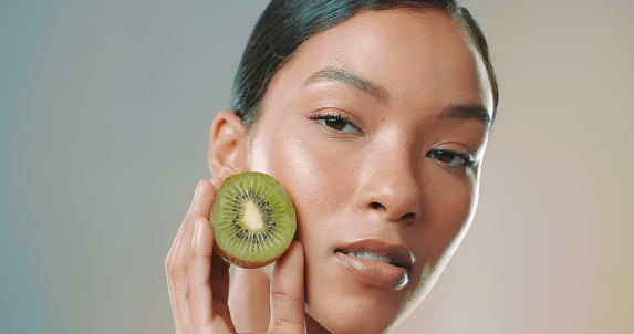 Close-up portrait of beautiful young woman with glowing face holding halved kiwi against gray background